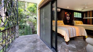 Un balcón con barandilla de hierro forjado, una cama con cubrecama estampado y un ventanal con vistas a la vegetación.