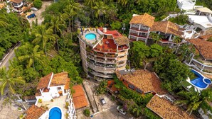 Un edificio de varios pisos con piscina en la azotea, rodeado de abundante vegetación y otras edificaciones residenciales.