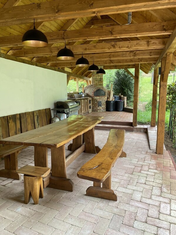 Un patio al aire libre con mesa y bancos de madera, un horno de ladrillo y luces colgantes.