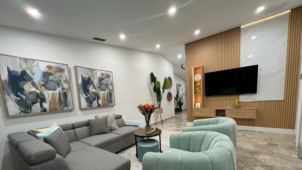 A modern living room with a sectional sofa, a teal armchair, a coffee table, and a flat-screen TV mounted on a wooden wall. - Flamingo  Dream Escape / 5 Bedrooms/ Htd Pool/ Game Room/ BBQ / Baby items (Miami)
