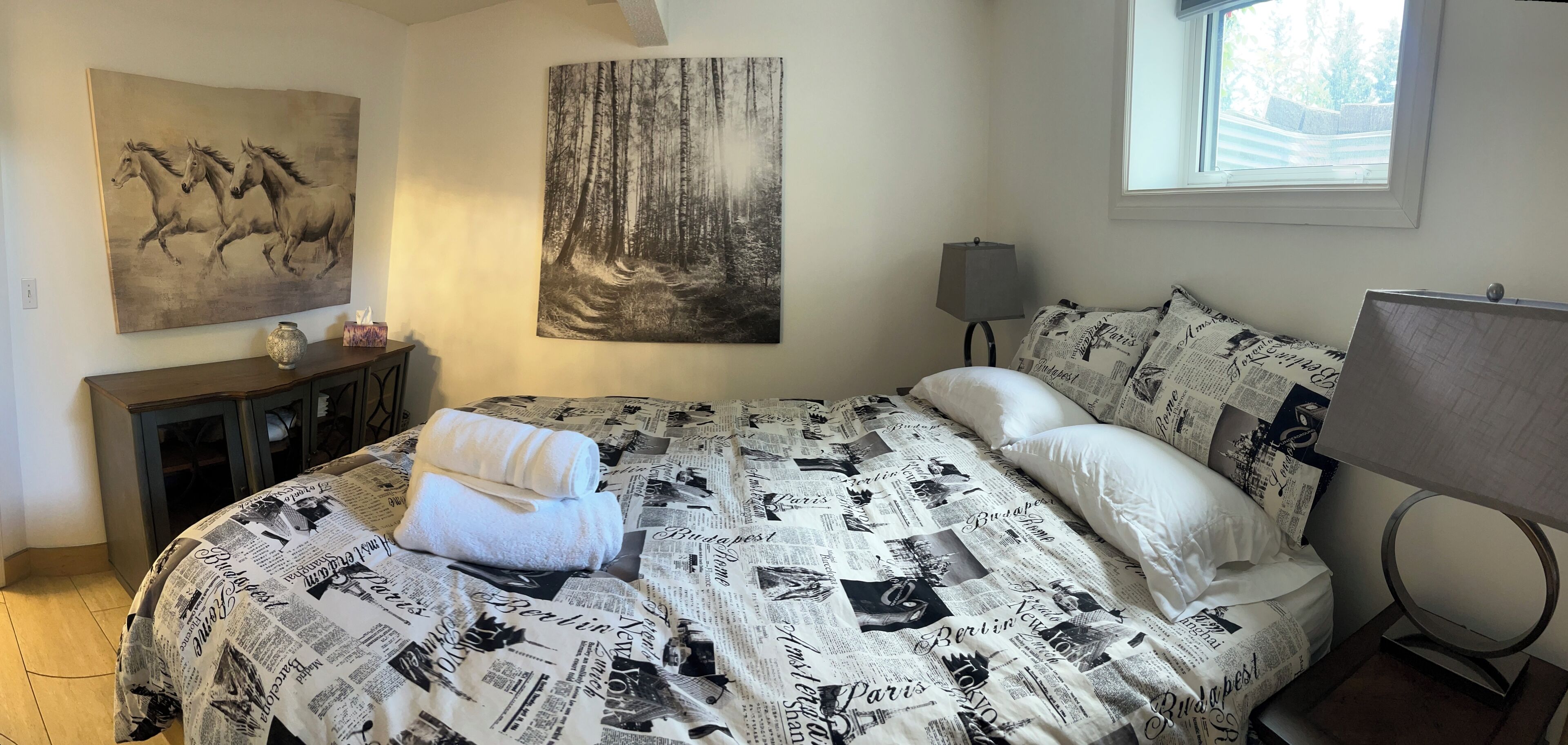 A bedroom with a bed covered in newspaper print, a large forest painting, a vase, and a lamp.