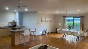 A modern kitchen with a central island, a dining area with white chairs, and a large window offering a view of greenery.
