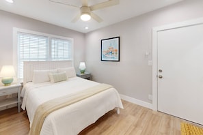 A bedroom with a bed, bedside tables, a ceiling fan, a window with blinds, a door, and a framed picture on the wall. - Walk to Beach & Johns Pass Village! Heated Pool ~ Private Patios ~ Free Parking & Amenities (Madeira Beach)
