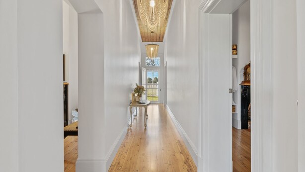 Un corridoio con pavimento in legno, pareti bianche, una lampada decorativa a soffitto e vista su un giardino attraverso una porta in vetro.