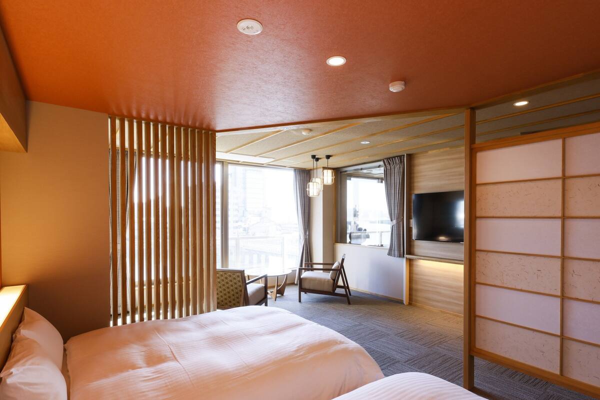 A hotel room with a large bed, a seating area with a chair, a TV mounted on the wall, and a view of the city through the window.
