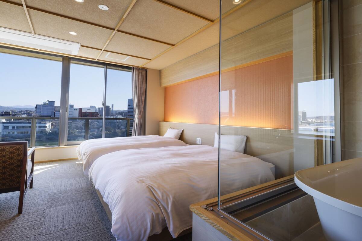 A hotel room with two beds, a large window with a city view, and a glass partition.
