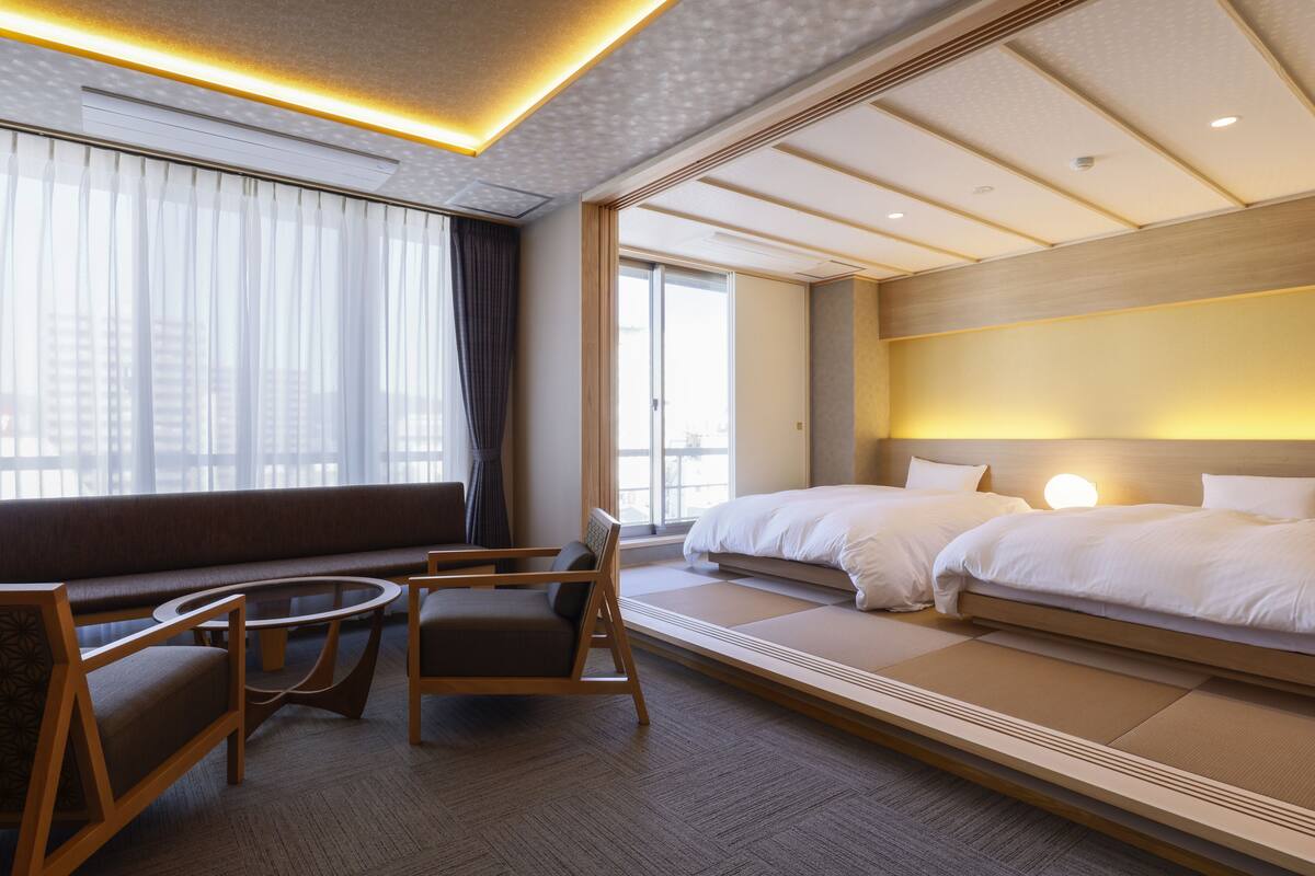 A hotel room with a sofa, two beds, a round table, and a large window with curtains.