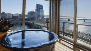 A room with a large window overlooking a cityscape and a bridge, featuring a hot tub and a wooden floor. - Ohashikan (Matsue)
