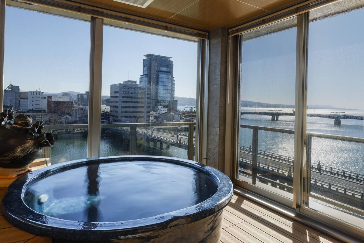 A room with a large window overlooking a cityscape and a bridge, featuring a hot tub and a wooden floor.