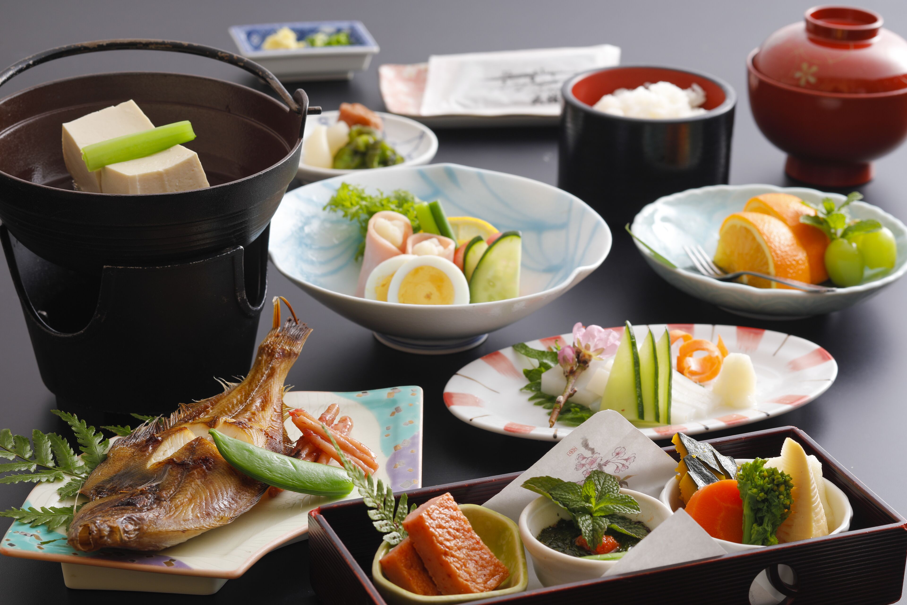 A variety of dishes including fish, vegetables, and tofu, arranged on a table with traditional Japanese bowls and a teapot.
