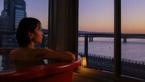 A person relaxing in a red bathtub by a large window overlooking a river and a bridge at dusk. - Ohashikan (Matsue)