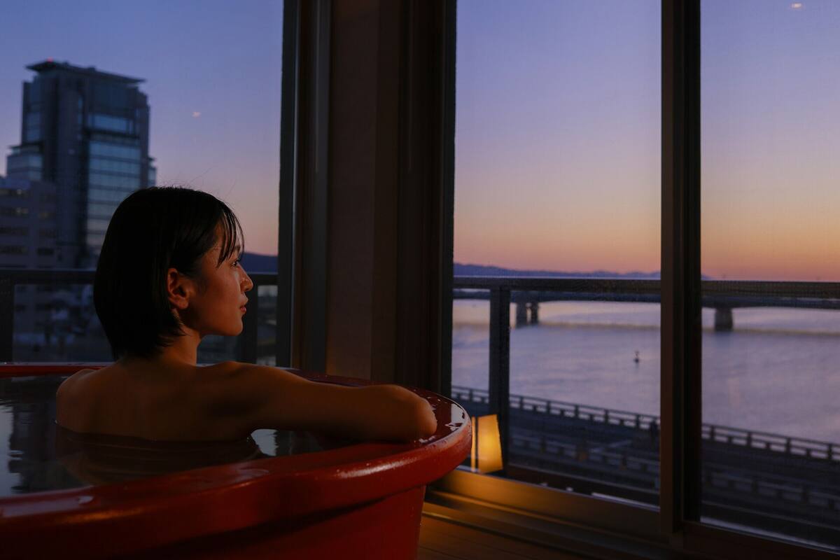 A person relaxing in a red bathtub by a large window overlooking a river and a bridge at dusk.