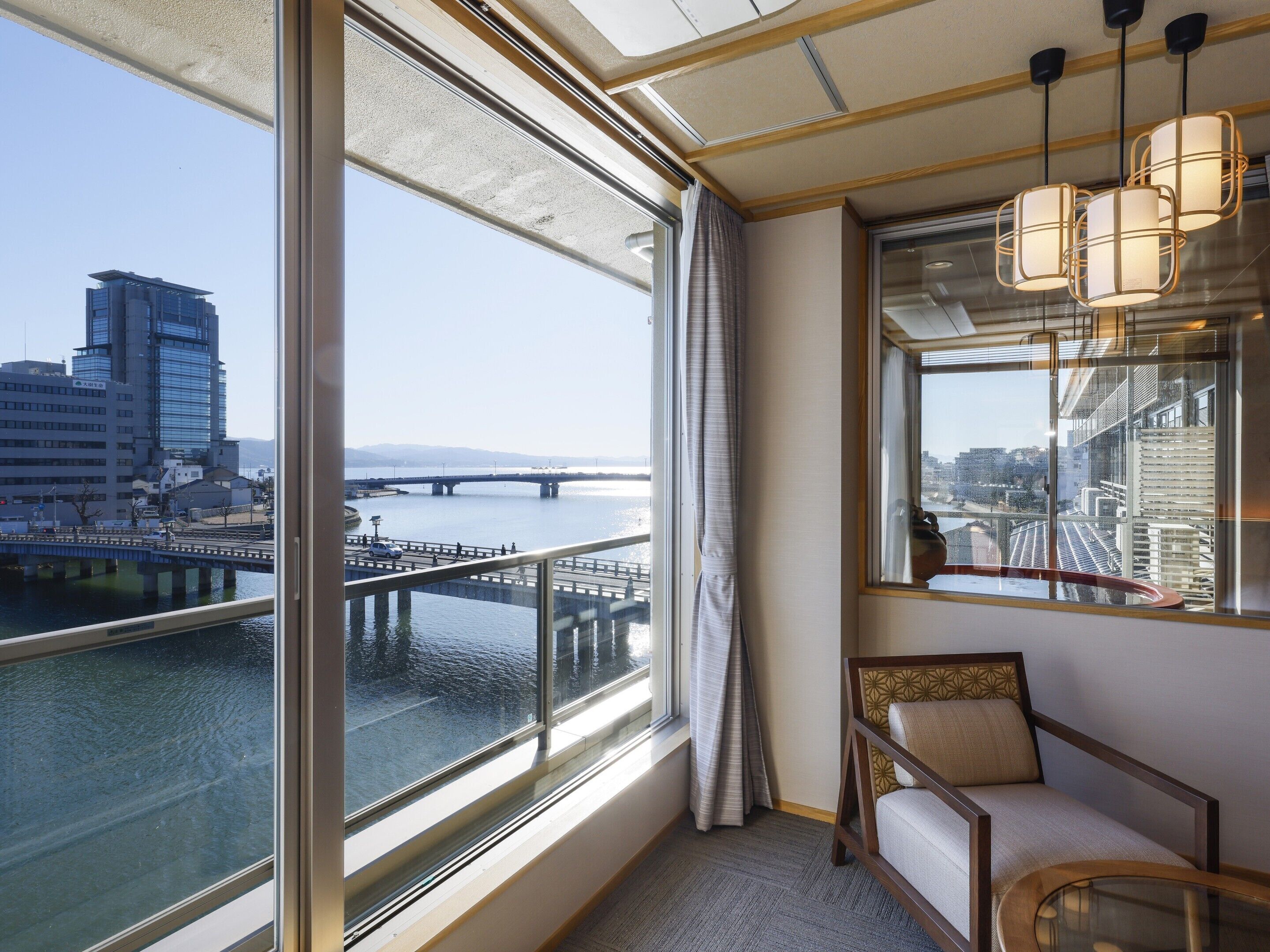 Habitación de hotel con un ventanal grande que ofrece vistas de un paisaje urbano, una silla de madera y una lámpara colgante.