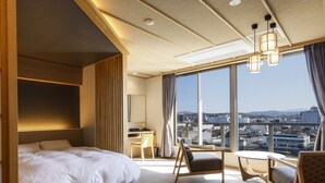 A modern hotel room with a large bed, a desk, a chair, and a view of the city through large windows. - Ohashikan (Matsue)