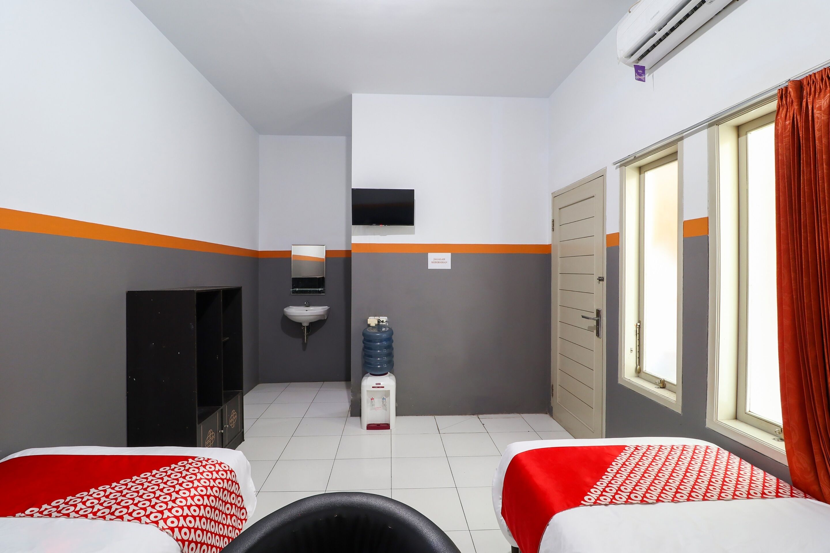 Habitación de hotel con dos camas, televisión, lavamanos, dispensador de agua y ventana con cortinas.