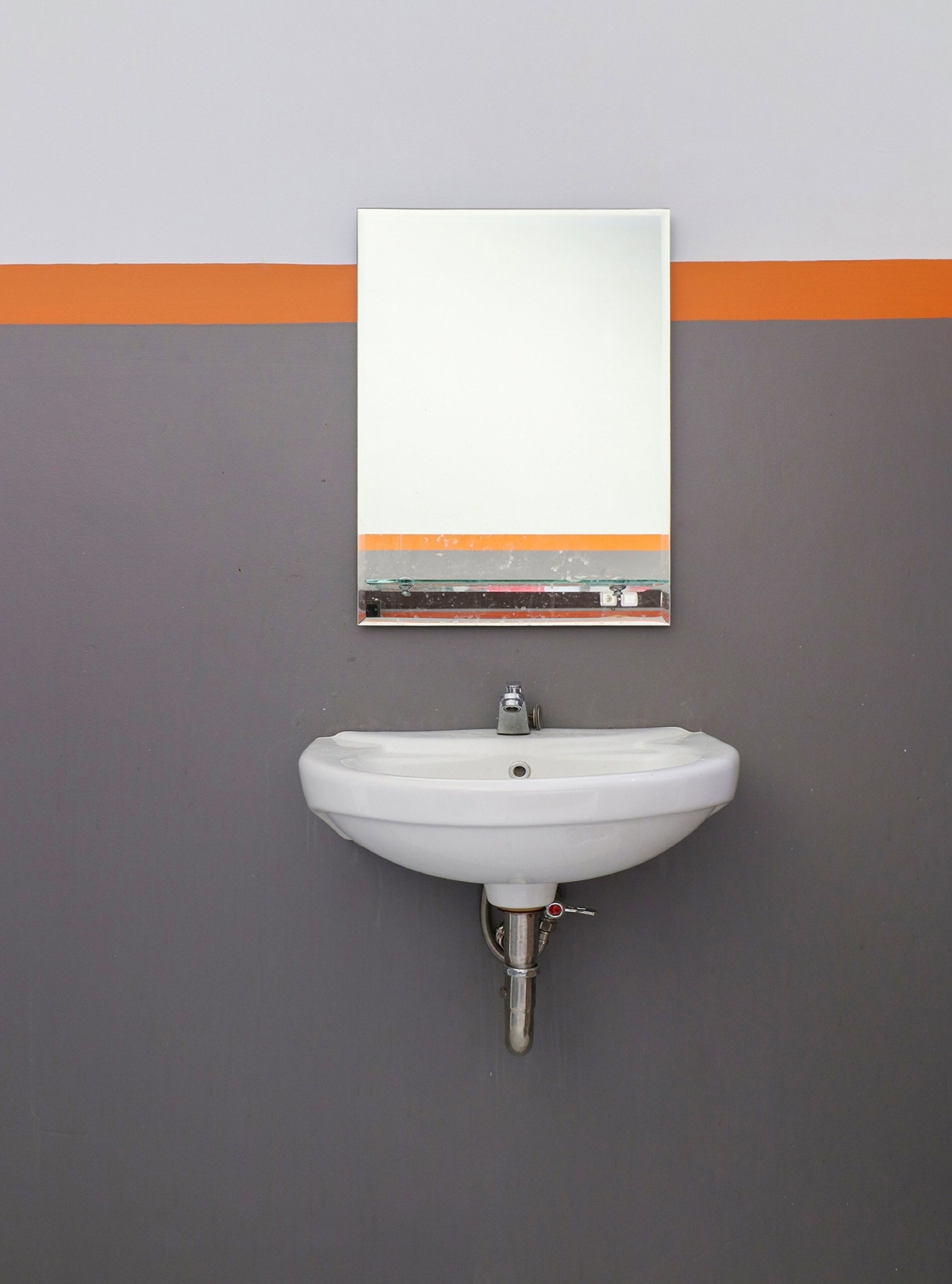 Een badkamer met een wit wastafel, een spiegel erboven en een grijze muur met een oranje streep.