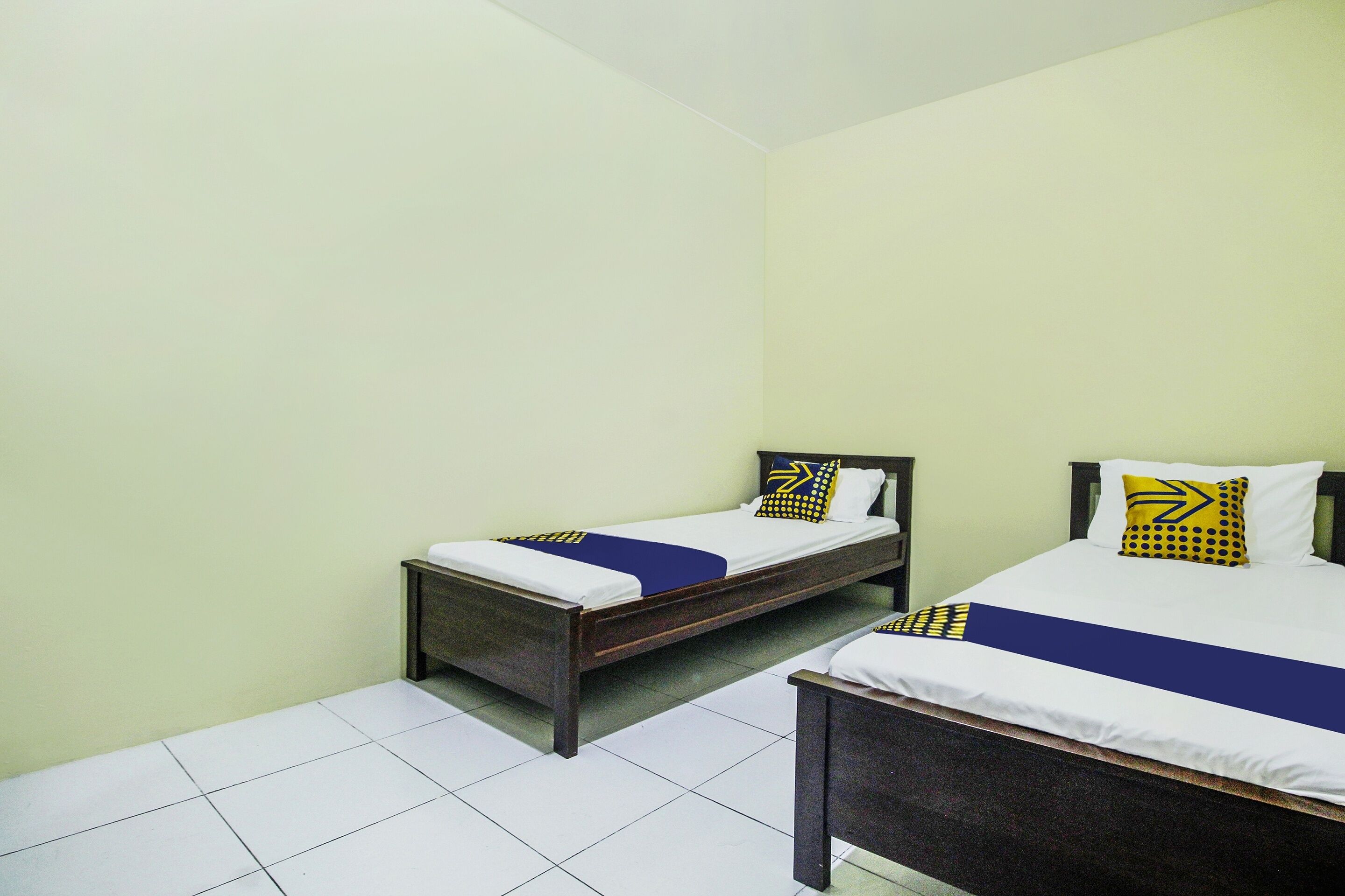A room with two single beds, each with a wooden frame and white bedding. The walls are painted light green, and the floor is tiled with white tiles.