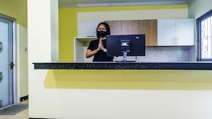 A reception desk with a computer monitor, a person standing behind it, and a yellow wall. - OYO Life 91606 Nadin Guest House Syariah (Garut)