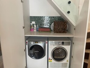 Un lavadero pequeño con dos lavadoras, un estante con productos de limpieza y un revestimiento de azulejos.