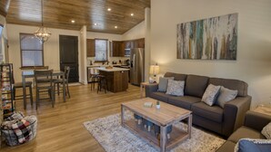 Een ruime woonkamer met een houten plafond, een grijze bank, een bijzettafeltje en een schilderij aan de muur.