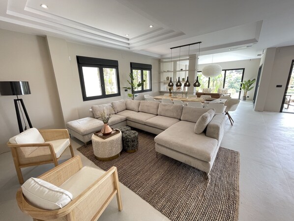 Una sala de estar amplia con un sofá seccional grande, un comedor con una mesa larga y una cocina al fondo.