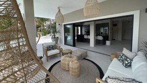 Ein modernes Wohnzimmer mit einer Couch, Sesseln und einem Korbstuhl. Zwei Pendelleuchten und ein großes Fenster sind vorhanden.
