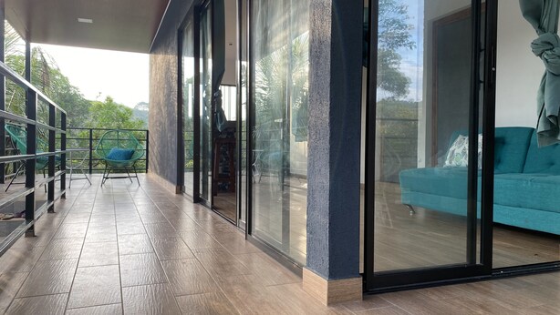 A modern living room with a teal sofa, a glass door leading to an outdoor area with chairs and a table, and a wooden floor. - Casa Melodía 🌿 2BR Near El Cuco & Las Flores (Chirilagua)
