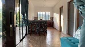 A modern kitchen with a stone backsplash, wooden bar stools, and a large window with vertical blinds. - Casa Melodía 🌿 2BR Near El Cuco & Las Flores (Chirilagua)