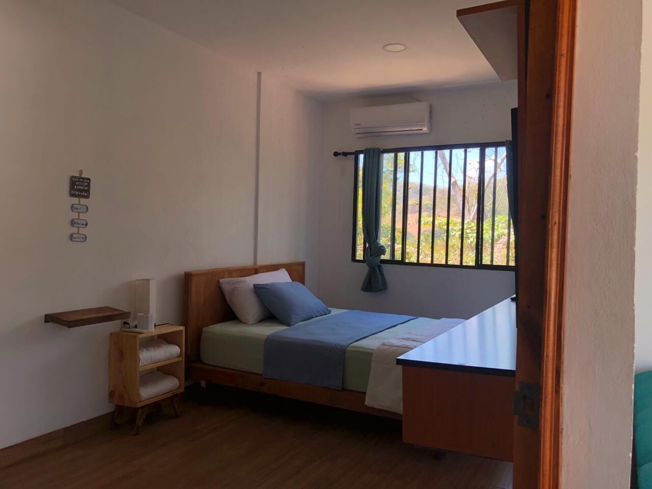 A compact bedroom with a bed, a small wooden shelf, a bedside table, and a window with a view.