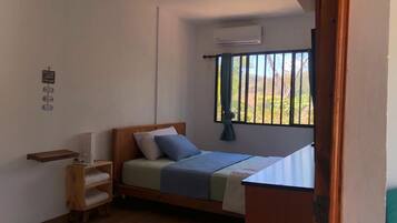A compact bedroom with a bed, a small wooden shelf, a bedside table, and a window with a view.