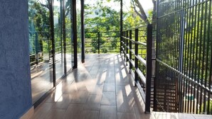 A corridor with glass walls and a wooden floor, leading to a balcony with a metal railing and a view of greenery. - Casa Melodía 🌿 2BR Near El Cuco & Las Flores (Chirilagua)