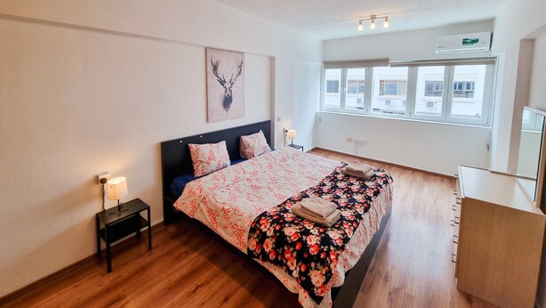 A bedroom with a bed, a nightstand, a lamp, a window, and a wall-mounted air conditioner.