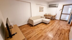 A living room with a wooden floor, a white sofa, a dining table with chairs, and a flat-screen TV on a wooden stand.