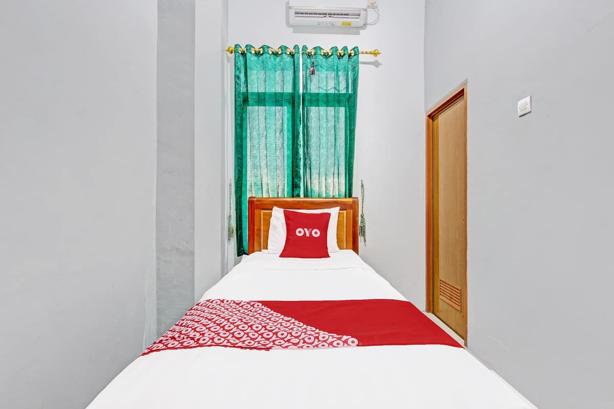 Une chambre d’hôtel étroite, avec un lit simple, une tête de lit en bois, un oreiller rouge et un rideau vert.
