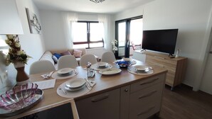 A dining area with a table set for four, a television on a stand, and a sofa in the background.