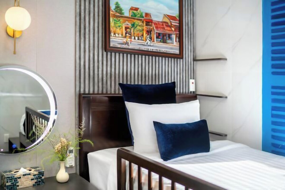 A hotel room with a bed, a round mirror, a framed picture on the wall, and a bedside table with a vase and a box.
