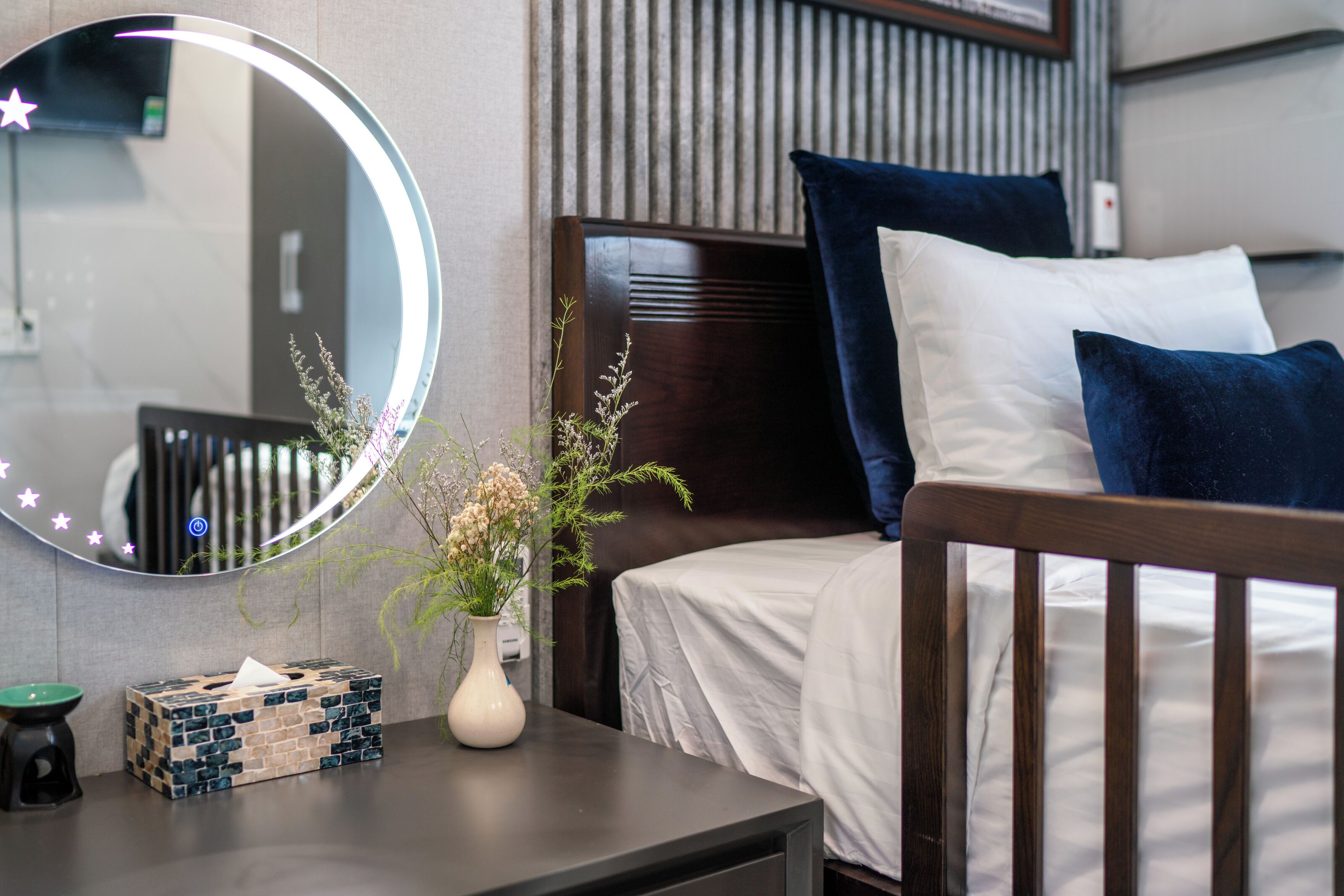 A neatly made bed with white and dark blue pillows, a bedside table with a vase of flowers, a mirror with a star light, and a small box of tissues.