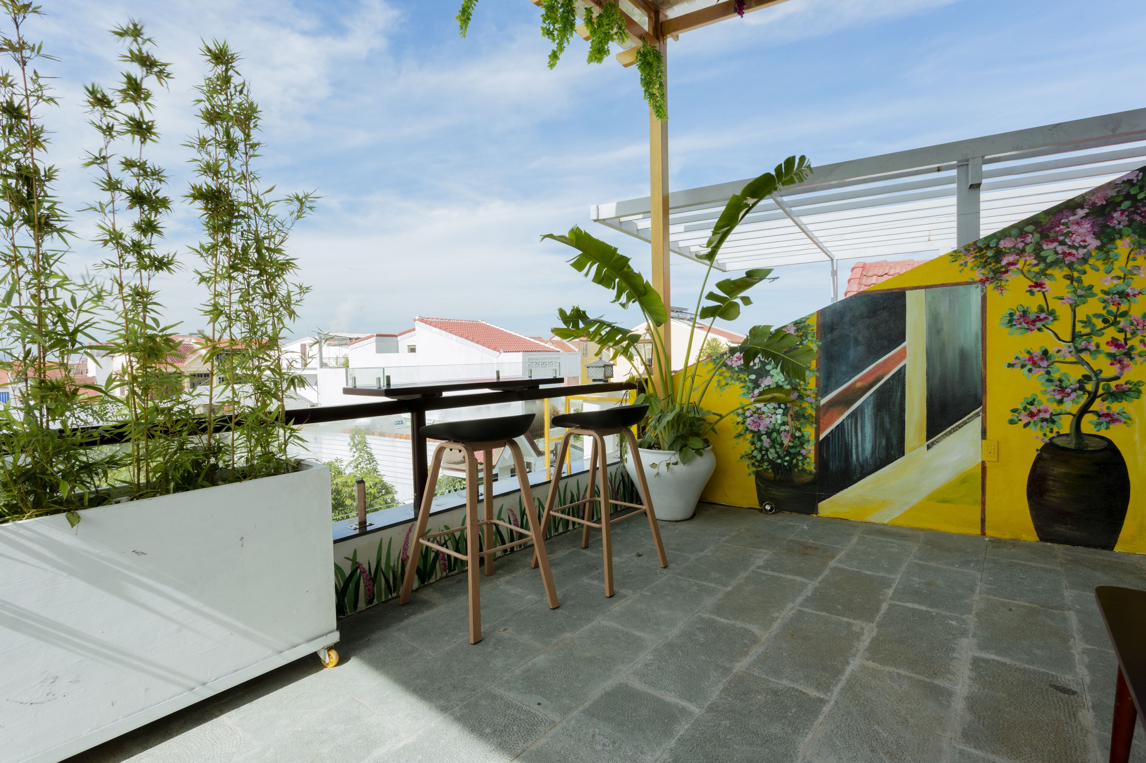 A balcony with a table and two chairs, potted plants, and a yellow wall with a floral mural.