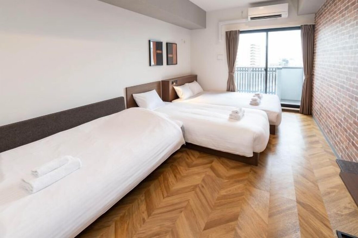A hotel room with two beds, a wooden floor, a brick wall, and a window with curtains.