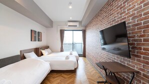 A hotel room with two beds, a TV mounted on a brick wall, a small table, and a balcony with curtains.