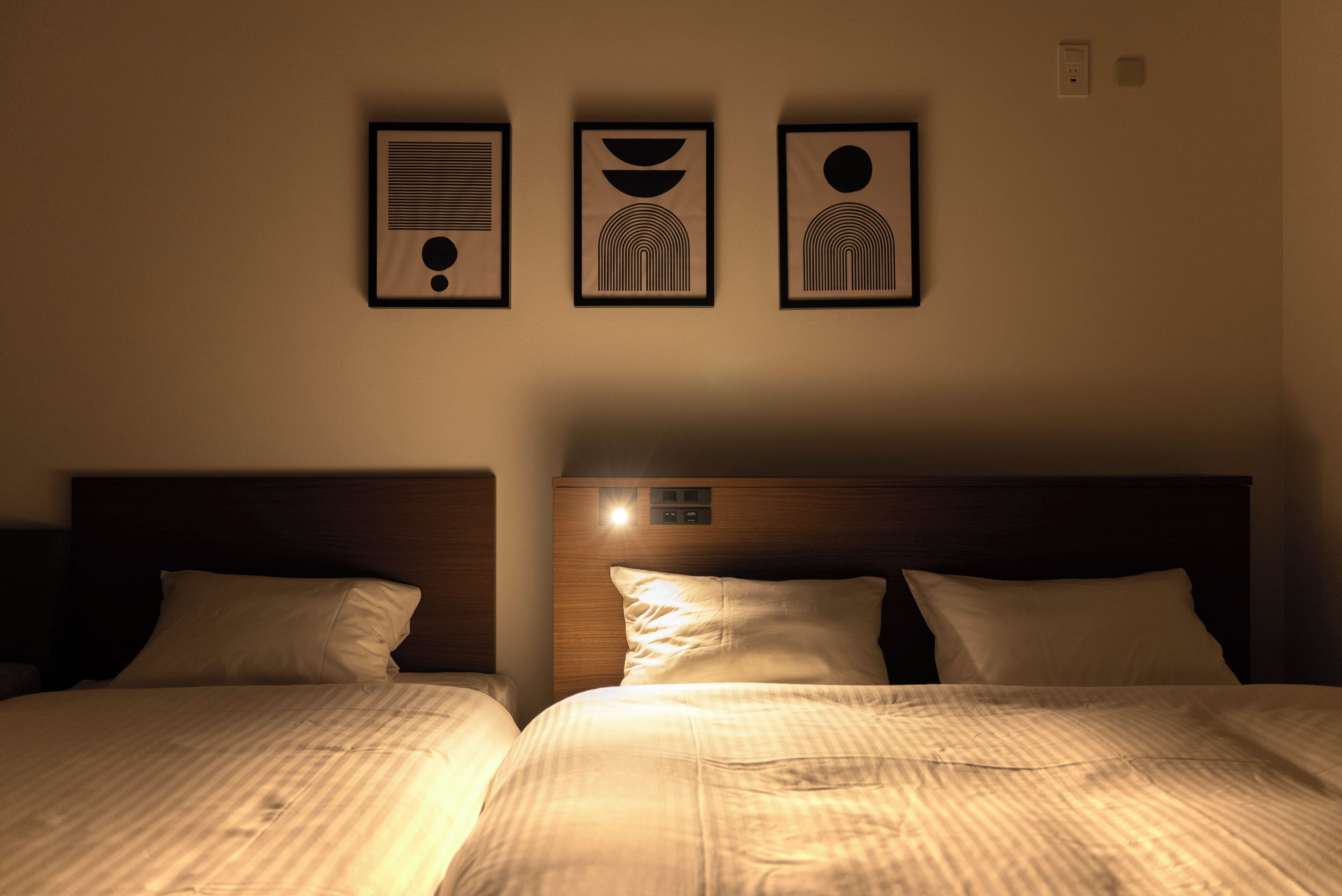 A hotel room with two beds, a headboard with a control panel, and three framed wall art pieces above the beds.