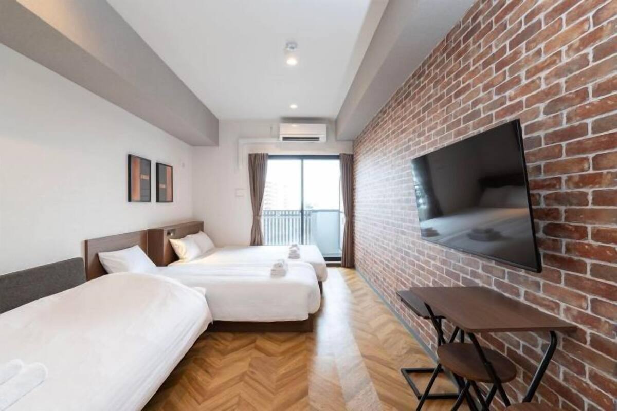 A hotel room with three beds, a TV mounted on a brick wall, a balcony, and a wooden floor.