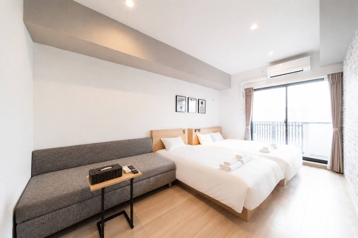 A modern hotel room with a sofa, two beds, a small table, and a balcony with curtains.