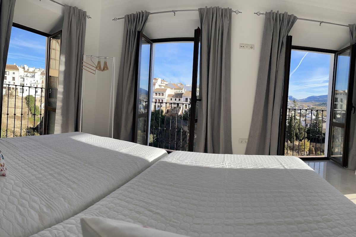 A bedroom with two beds, large windows with curtains, and a view of buildings and mountains.
