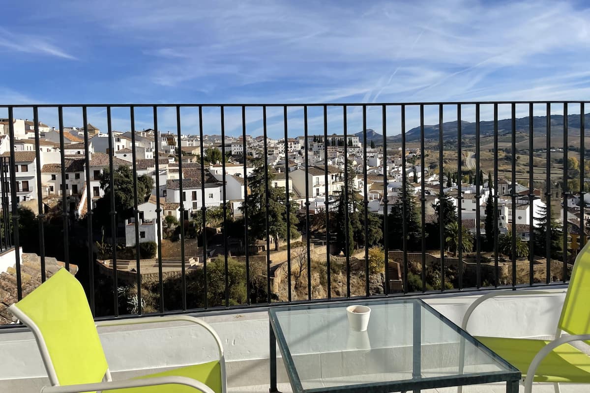 A balcony with a glass table and two chairs, overlooking a town with white buildings and a clear blue sky.