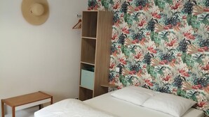 Ein Schlafzimmer mit Bett, einer Hängeleuchte, einem Kleiderschrank, einem kleinen Tisch und einer Wand mit tropischem Blattmuster.