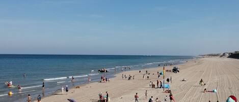 A wide sandy beach with people walking and sunbathing, a clear blue sky, and the ocean in the background.