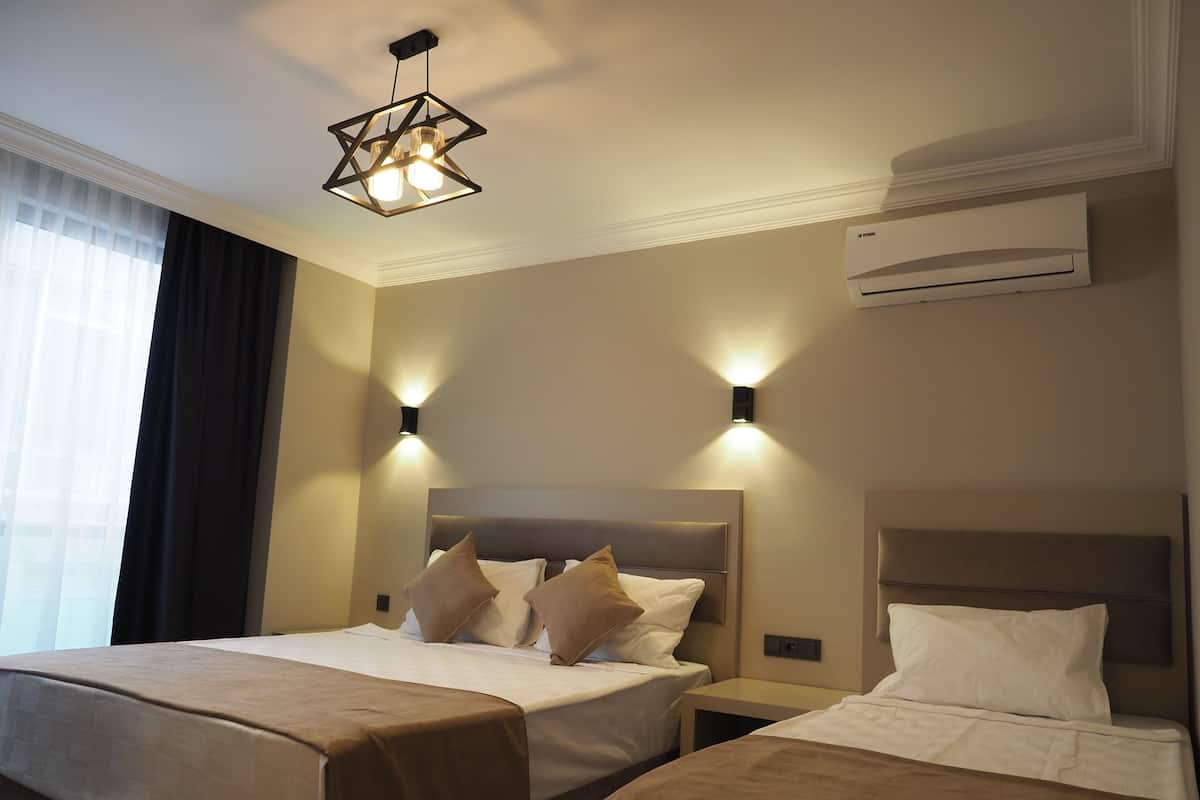 A hotel room with two beds, a modern chandelier, wall-mounted lights, an air conditioning unit, and a large window with curtains.