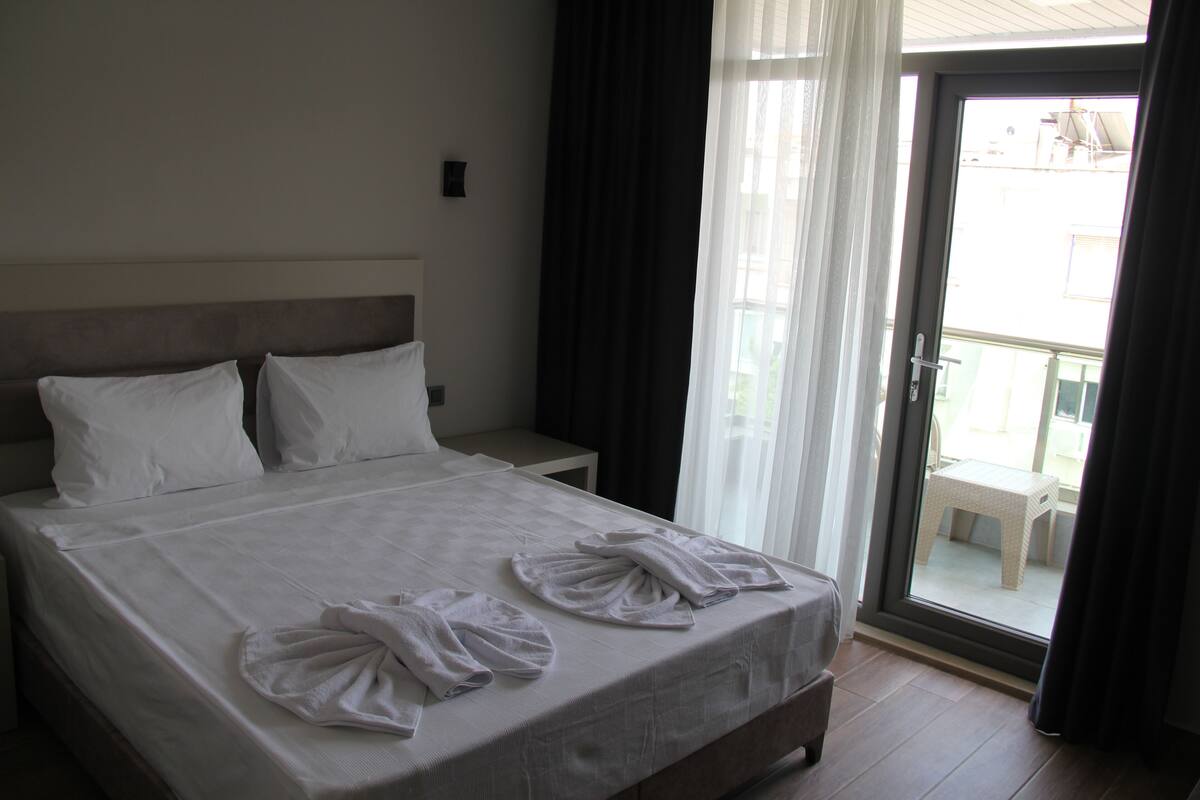 A hotel room with a bed, a nightstand, a window with curtains, and a view of the outside.