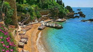 A beach with clear blue water, a stone building with a red roof, and a staircase leading down to the beach. - Zenia Otel (Antalya)
